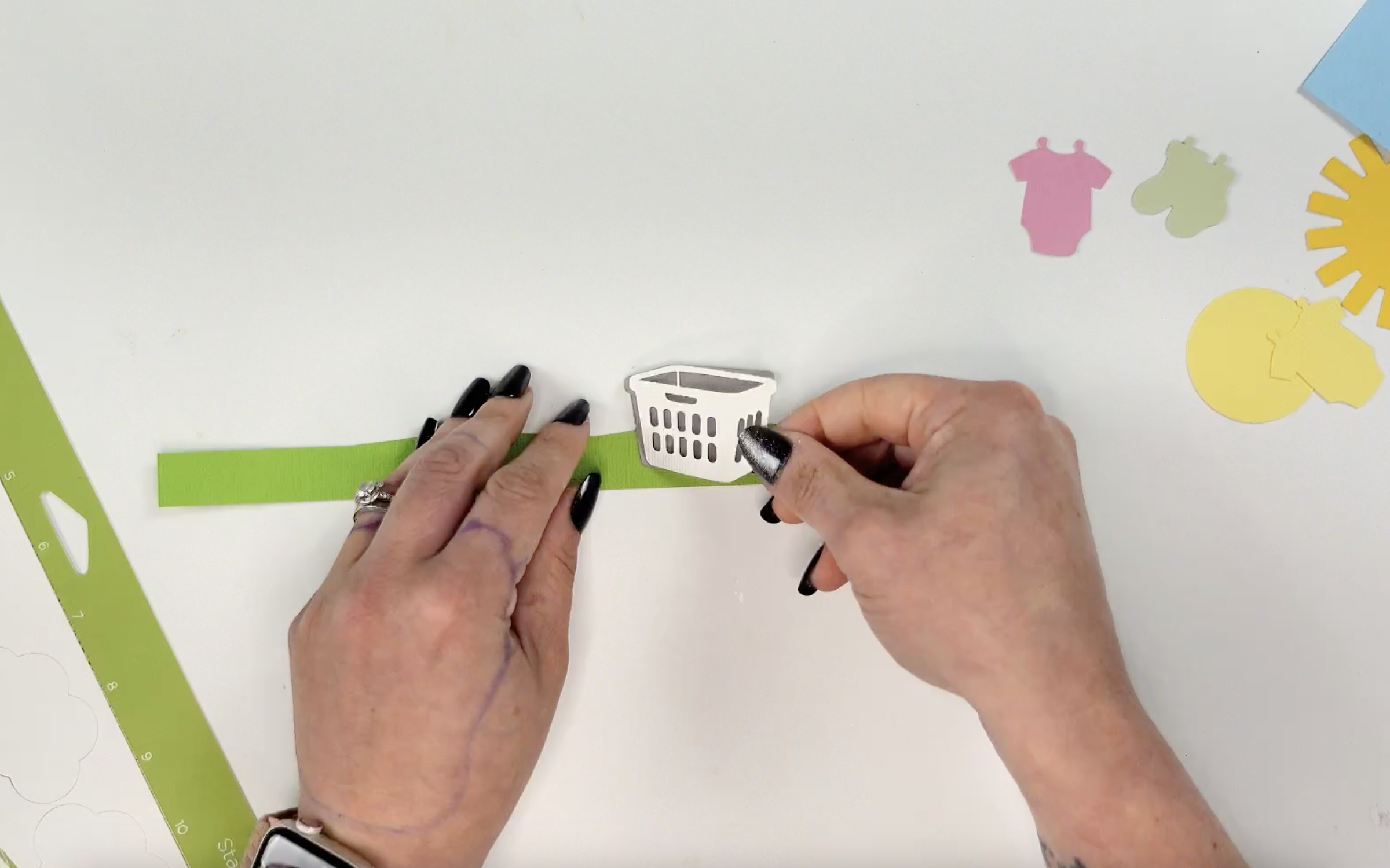 Close-up of hands assembling a green cardstock strip with a layered white baby laundry basket. Additional cut pieces like a pink onesie, yellow sun, and green baby clothes are visible nearby.